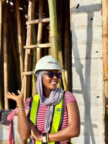 Saida in a hard hat and hi-vis, smiling beside a bamboo scaffold